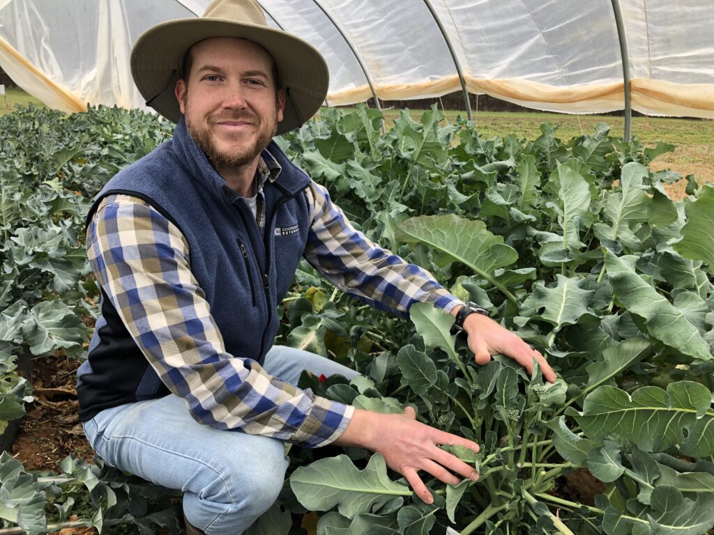 A man in a wide brimmed hat displays his plants in a high tunnel.