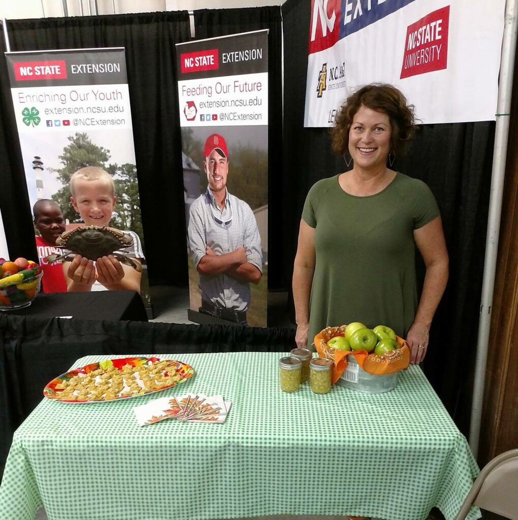 A woman at a conference standing behind a display of apples and apple products.